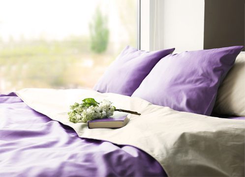 ordentlich gemachtes Bett mit lila Laken und Kissen, vor einem Fenster mit Blick auf eine grüne Wiese
